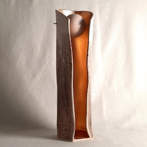 Peut inclure: Un vase en céramique rectangulaire et élancé, avec un extérieur texturé marron et blanc moucheté. L'intérieur est de couleur dorée chaude. Un objet arrondi de couleur claire est suspendu à l'intérieur. Le vase a une forme organique unique.