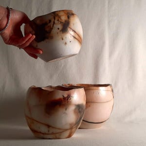 Puede incluir: Tres vasijas de cerámica con formas orgánicas y efecto marmolado. Las vasijas tienen una paleta de colores rosa claro y marrón. Una vasija es sostenida por una mano. Las vasijas son probablemente para uso decorativo o funcional.