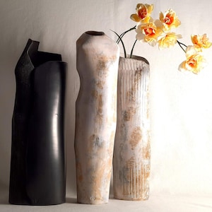 Peut inclure: Trois vases décoratifs de hauteurs et de formes variées. Le vase le plus à gauche est noir et cylindrique. Le vase du milieu est blanc cassé avec une surface texturée. Le vase de droite est beige avec des lignes verticales et contient des fleurs jaunes et oranges.