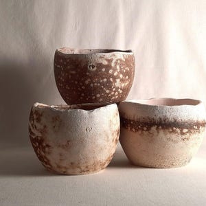 Puede incluir: Tres cuencos de cerámica hechos a mano en tonos marrones y blancos. Los cuencos tienen una superficie texturizada con un patrón moteado. Los cuencos están apilados, con el cuenco más grande en la parte superior. Los cuencos probablemente se utilizan para servir alimentos o como objetos decorativos.