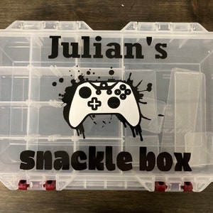 May include: A clear plastic snack box with multiple compartments. The box features the name "Julian's" above a black and white controller graphic and the words "snackle box" below. Red clasps secure the lid.