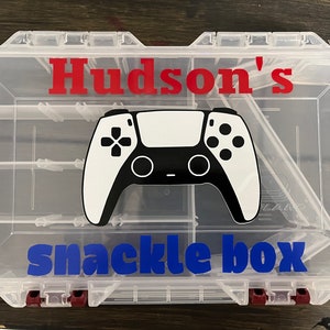 May include: A clear plastic snack box with compartments, featuring a black and white video game controller graphic. The box has the name "Hudson's" in red and "snackle box" in blue.