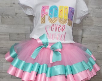 May include: A pink tutu with teal ribbon trim and a white t-shirt with the text "Four ever sweet" and a colorful ice cream cone graphic.