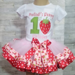 Pu&ograve; includere: Una maglietta bianca con il testo "Melodi's 1st Birthday" e un numero 1 verde con una fragola rossa in cima. La maglietta &egrave; abbinata a una gonna di tulle rosa con un nastro a pois rossi e bianchi e un nastro con stampa di fragole.