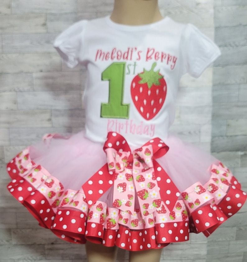 Pu&ograve; includere: Una maglietta bianca con il testo "Melodi's 1st Birthday" e un numero 1 verde con una fragola rossa in cima. La maglietta &egrave; abbinata a una gonna di tulle rosa con un nastro a pois rossi e bianchi e un nastro con stampa di fragole.