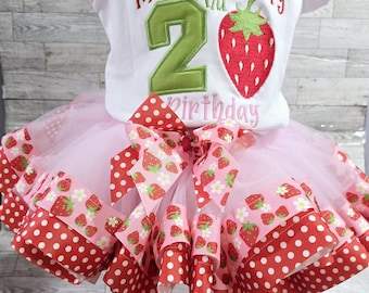 May include: A pink tulle tutu with red and white polka dot ribbon and a strawberry print ribbon. The tutu is paired with a white t-shirt with the text "Madeline's Berry 2nd Birthday" and a green number 2.