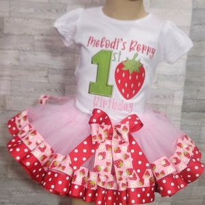 Pu&ograve; includere: Una maglietta bianca con il testo "Melodi's Berry 1st Birthday" e un numero 1 verde con una fragola rossa. La maglietta &egrave; abbinata a una gonna di tulle rosa con un nastro a pois rossi e bianchi e un nastro con stampa di fragole.