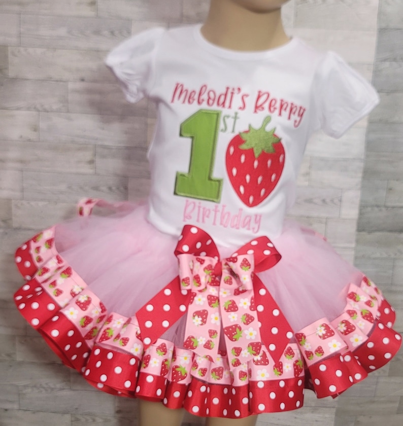 Pu&ograve; includere: Una maglietta bianca con il testo "Melodi's Berry 1st Birthday" e un numero 1 verde con una fragola rossa. La maglietta &egrave; abbinata a una gonna di tulle rosa con un nastro a pois rossi e bianchi e un nastro con stampa di fragole.
