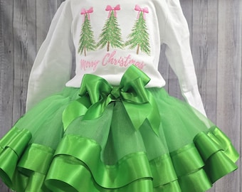 May include: A white long-sleeve shirt with a Christmas tree design and the text "Merry Christmas". The shirt is paired with a green tutu skirt with a large bow. The skirt has layers of tulle and satin ribbon.
