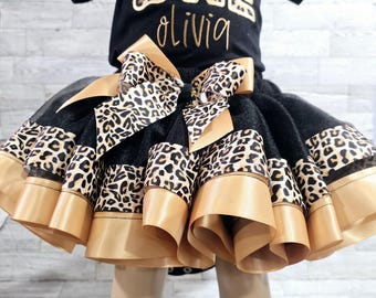 Puede incluir: Una falda de tul negra con ribetes de cinta dorada y estampado de leopardo. La falda la lleva puesta un niño y tiene un gran lazo dorado en la parte delantera. La falda es perfecta para una fiesta de primer cumpleaños.