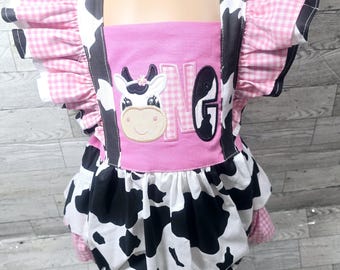 May include: A pink and white cow-themed romper with a cow face applique and the number "1" and the letter "G". The romper features black and white cow print fabric, pink gingham ruffle sleeves, and black straps.