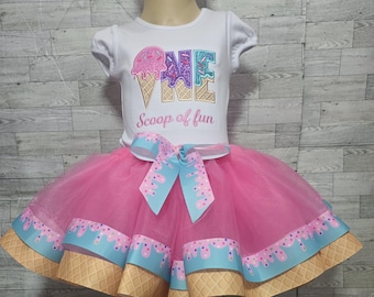 May include: A white short-sleeved shirt with a pink, purple, and blue ice cream cone design that spells out "ONE" and the text "Scoop of fun". The shirt is paired with a pink tulle skirt with a blue and white ice cream cone design.