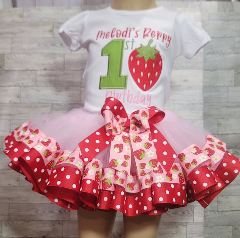 Pu&ograve; includere: Un tut&ugrave; di tulle rosa con nastro a pois rossi e bianchi e nastro con stampa di fragole. Il tut&ugrave; &egrave; abbinato a una maglietta bianca con il testo "Melodi's Berry 1st Birthday" e un numero 1 verde con una fragola rossa.