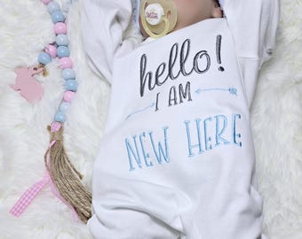 May include: A white baby onesie with blue and black embroidery that reads "hello! i am new here". The onesie is on a white fuzzy surface with a pink and blue beaded necklace and a pink bunny toy.