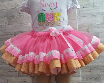 May include: A pink tulle tutu skirt with a white ribbon bow. The skirt has a layered design with a white and brown ice cream cone print. The skirt is decorated with white and pink icing and sprinkles. The text "Sweet One" is embroidered on the white shirt.
