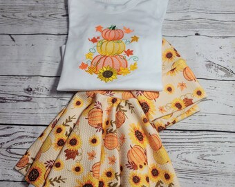 May include: A white sleeveless shirt with an embroidered design of three stacked pumpkins and sunflowers. The shirt is paired with a cream-colored skirt with a pumpkin and sunflower print.
