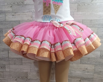May include: A pink tulle tutu skirt with a brown, white, and blue ice cream cone design. The skirt has a white underskirt and a brown ribbon bow. The skirt is for a child and has the name "Olivia" embroidered on the front.