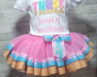 May include: A white short-sleeved shirt with the embroidered text THREE Scoops of Sweetness in pastel colors, paired with a pink tulle tutu skirt featuring a blue ribbon trim with a colorful sprinkle pattern and a waffle cone print border.