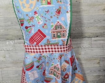 May include: A blue and red Christmas-themed apron with a gingerbread house and cookie design. The apron has a heart-shaped top and a ruffled skirt. The text "Christmas Cookies" is printed on the apron.
