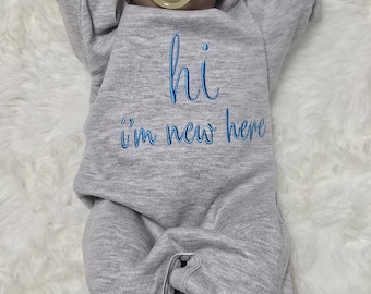 May include: A newborn baby doll wearing a gray hooded onesie with the words "hi i'm new here" embroidered in blue. The doll has a pacifier that says "Love Mom". The doll is lying on a white fluffy surface.