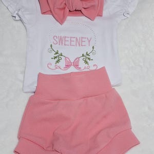 Può includere: Un completo per neonati composto da una maglietta bianca a maniche corte con il nome "SWEENEY" ricamato in verde e rosa, una fascia rosa con fiocco e pantaloncini rosa abbinati. La maglietta presenta un fiocco decorativo e un motivo floreale.