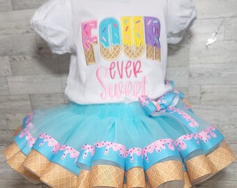 May include: A white short-sleeved t-shirt with the text FOUR EVER SWEET in colorful ice cream cone lettering, paired with a light blue tulle tutu skirt featuring a ribbon trim printed with a pink icing and waffle cone pattern.
