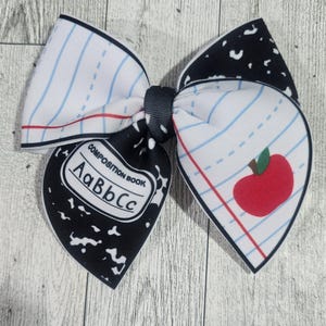 May include: A black and white composition book bow with a red apple on a white and blue lined background. The bow is tied with a black ribbon.