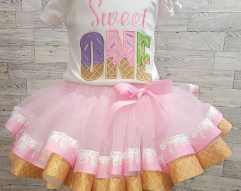 May include: A white short-sleeved bodysuit with the embroidered text Sweet ONE in pink, purple, and green ice cream cone lettering, paired with a layered light pink tulle tutu skirt featuring a ribbon trim with an ice cream cone pattern.