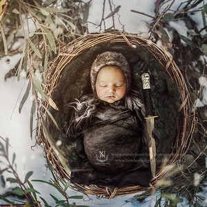 May include: A newborn baby is sleeping in a woven basket, wrapped in a dark brown fabric with a hood. The baby is surrounded by green foliage and a small silver sword is lying next to them.