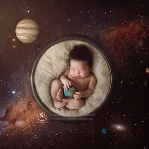May include: A newborn baby is sleeping in a white, round, fluffy nest. The baby is holding a small, blue and green globe. The background is a dark blue space with stars and planets.