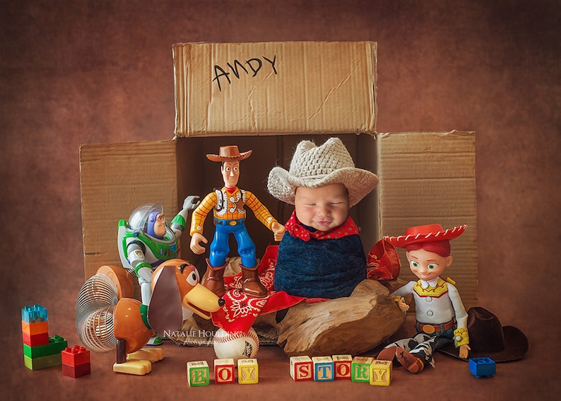 May include: A newborn baby wearing a cowboy hat and a red bandana is lying in a cardboard box with the word "ANDY" written on it. The baby is surrounded by toys from the Toy Story movie, including Woody, Buzz Lightyear, Slinky Dog, and a set of blocks that spell out "BOY STORY".
