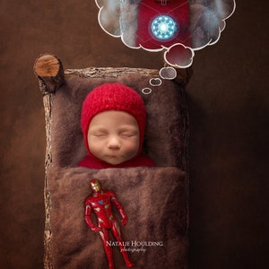 Puede incluir: Un bebé con un gorro rojo duerme en una cuna de madera con un juguete de Iron Man rojo a su lado. Una burbuja de pensamiento sobre la cabeza del bebé muestra a Iron Man en una pose de superhéroe. El texto "NATALIE HOULDING Photography" está en la parte inferior de la imagen.