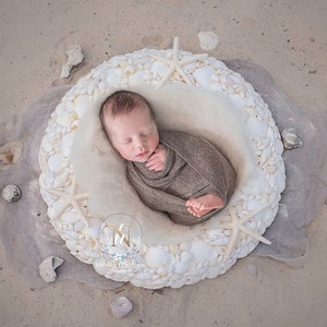 May include: A newborn baby is sleeping in a white seashell-shaped nest. The nest is filled with white seashells and the baby is wrapped in a brown blanket.