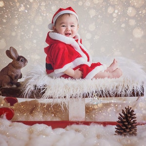 Può includere: Un bambino vestito con un costume rosso di Babbo Natale è seduto su una slitta ricoperta di pelliccia bianca con una statuetta di coniglio marrone accanto. Lo sfondo è marrone chiaro con un effetto nevoso.