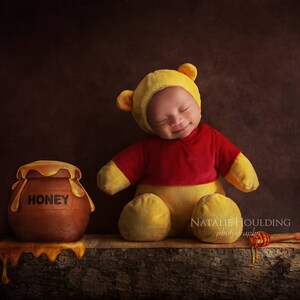 Puede incluir: Un bebé vestido con un disfraz de Winnie the Pooh, con una camisa roja y pantalones amarillos, está durmiendo con un tarro de miel y un cucharón de miel sobre una superficie de madera. El tarro de miel está etiquetado con "HONEY".