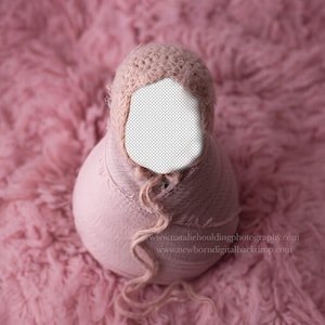 May include: A pink crocheted baby hat with a long, knotted tie. The hat is on a pink fluffy surface.