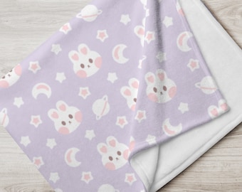 Può includere: Una morbida coperta in pile di colore lilla con un motivo ripetitivo di coniglietti bianchi, stelle, pianeti e lune crescenti. La coperta è piegata a metà, mostrando il retro in pile bianco.