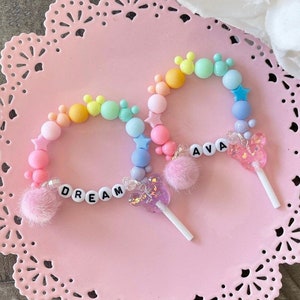 May include: Two colorful beaded bracelets with charms. One bracelet has the word "DREAM" spelled out in white letters and a pink butterfly charm. The other bracelet has the name "AVA" spelled out in white letters and a pink Mickey Mouse charm.