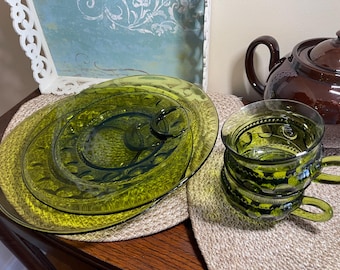 May include: A set of vintage green glass dishes and a teacup with a matching saucer. The teacup has a handle and is stacked. A brown teapot is in the background. The dishes are on a woven placemat.