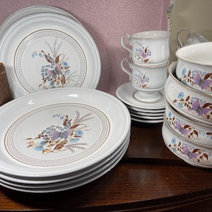 May include: A collection of white ceramic dinnerware featuring a central floral and butterfly motif in shades of blue, purple, and brown. The set includes stacked dinner plates, a stack of bowls, and two teacups with saucers, all decorated with a repeating geometric border pattern.