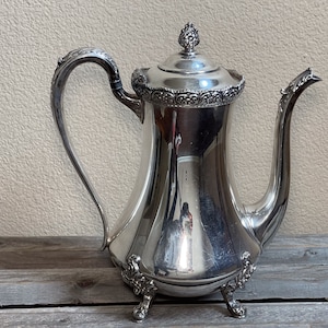 May include: A silver teapot with an ornate design and a decorative lid. The teapot has a curved body and a spout with a decorative handle. The teapot is sitting on a wooden surface.