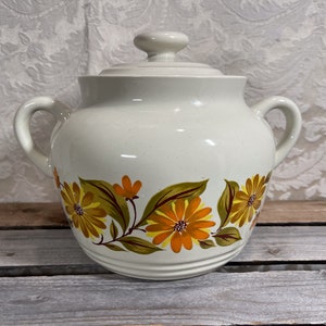 May include: White ceramic crock with a lid, featuring a floral design of yellow and orange flowers with green leaves around the body.