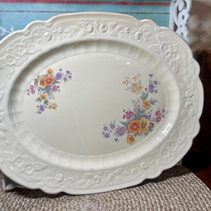 May include: Off-white oval serving platter with a scalloped edge and floral detailing. The platter features clusters of orange, yellow, and purple flowers. The rim has embossed floral and geometric patterns.