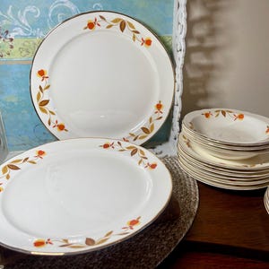 May include: A collection of white ceramic plates and bowls with a gold rim and a decorative floral pattern in orange, yellow, and brown. The plates are arranged on a dark wooden surface.