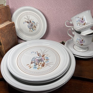 May include: A set of white ceramic dinnerware featuring a central floral and butterfly motif in shades of blue, purple, and brown, surrounded by a circular geometric border. The collection includes stacked dinner plates, a side plate, and two teacups.