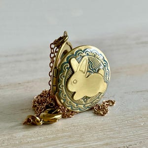 May include: A gold-coloured locket featuring a bunny design. The oval locket has a detailed border and a chain. The bunny is in the centre of the locket. The locket is open, revealing the inside.