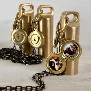 May include: Three gold-toned metal necklaces with different charms. The charms include a heart with the letter T, a heart with the letter K, and a round locket with a photo of two dogs. The necklaces are on a white surface.