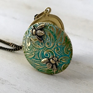May include: A round, antique-style pendant necklace with a teal and gold bee design. The pendant features two raised bee figures and intricate floral patterns. The necklace has a dark chain and a clasp.