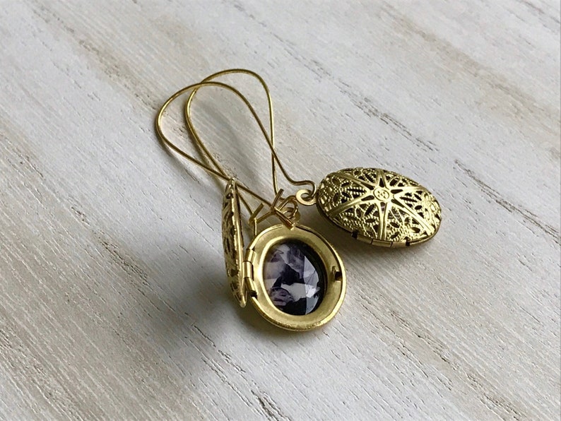 May include: Gold filigree locket earrings with a small, round, purple photo inside. The earrings are open and the back of the locket is visible.