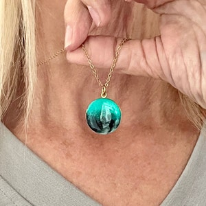 May include: A gold chain necklace with a round pendant featuring a teal and black abstract design.
